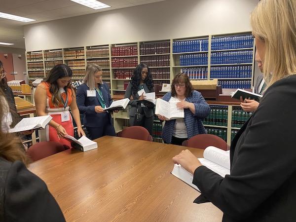 A law librarian standing at a table with a group of law students, showing them a section of a book she is holding up.